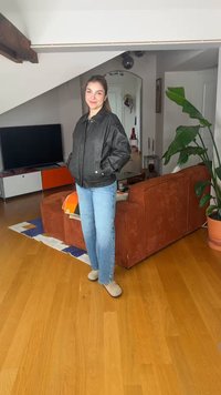 O tânără stă într-un living cu podea din lemn, canapea de culoare ruginie, o plantă mare verde în ghiveci și un televizor pe un dulap jos.