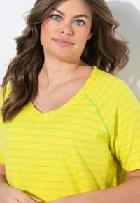 Haut jaune à manches courtes avec des rayures verticales vertes, présentant un décolleté en V et des détails de couture subtils, affiché sur un fond clair.