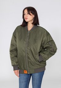 Veste bomber verte avec une finition brillante, poignets côtelés, deux poches zippées et une étiquette orange à l'ourlet. Dispose d'une fermeture éclair à l'avant.
