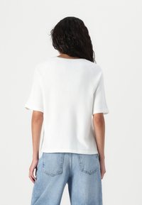 Maglione corto bianco a maniche corte con scollatura rotonda, caratterizzato da spacchi laterali e una consistenza morbida, abbinato a jeans azzurri a vita alta.