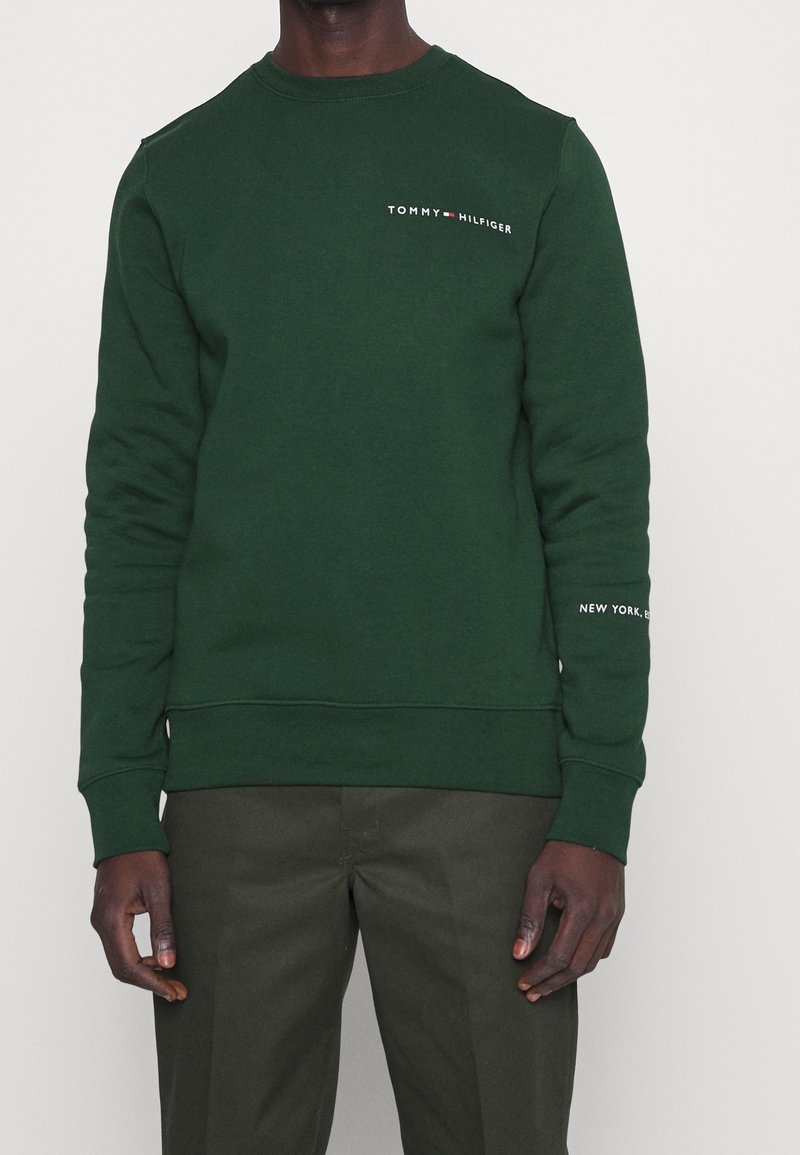 Sweat-shirt col rond vert foncé avec logo "Tommy Hilfiger" sur la poitrine et texte "New York, EST" sur la manche gauche, porté avec un pantalon vert olive.
