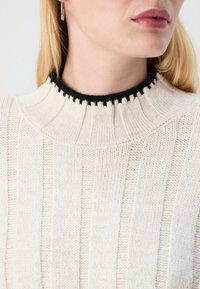 Ljus krämfärgad tröja med vertikalt ribbat mönster, med en svart scalloped krage. Närbilden visar detaljer kring halsringningen och tyget.