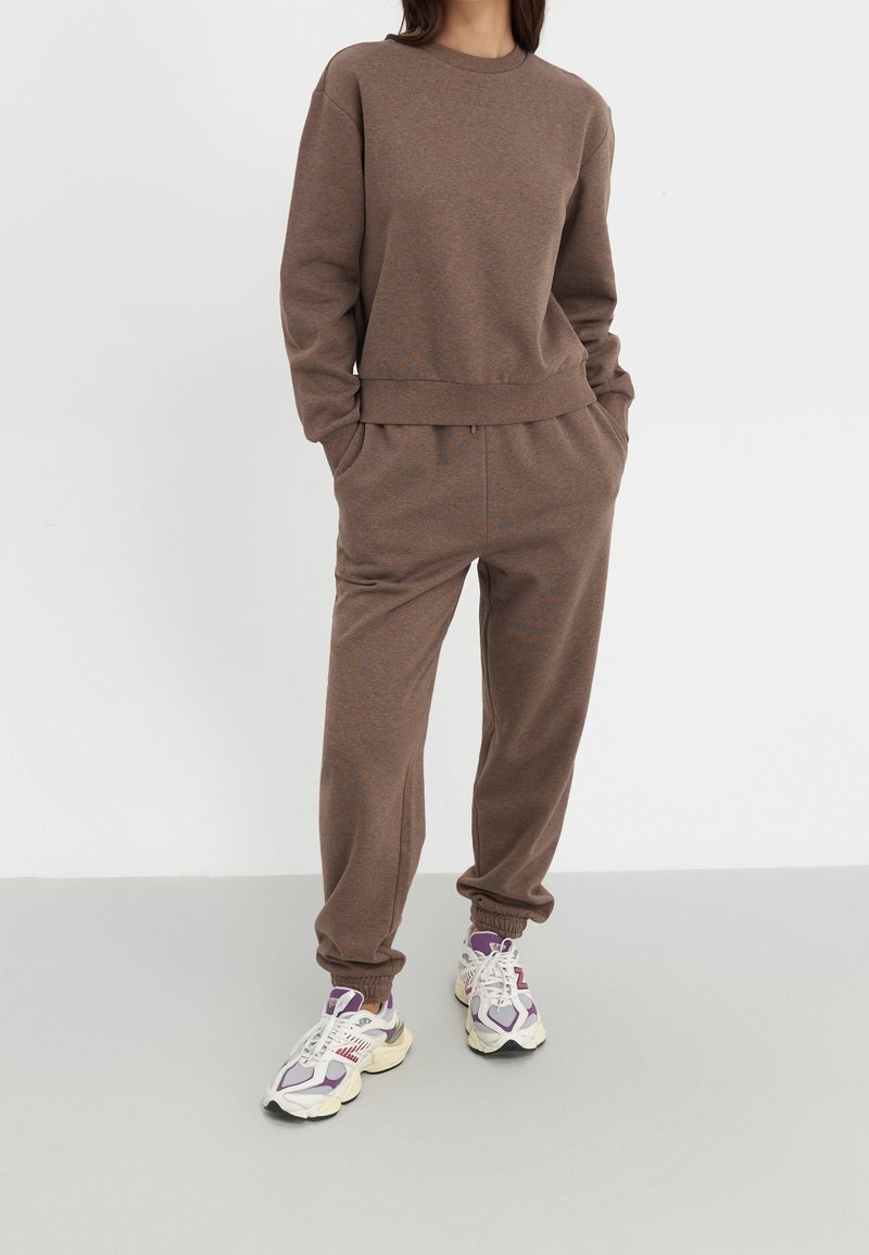 Personne portant un sweat-shirt à manches longues marron et un pantalon de jogging assorti, les mains dans les poches, chaussée de baskets blanches et violettes, debout.