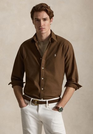 Junger Mann mit braunen Haaren, der ein braunes Hemd mit Knöpfen über einem beigen T-Shirt trägt, weiße Hose, brauner Gürtel und grüne Armbanduhr, Hände in den Taschen.