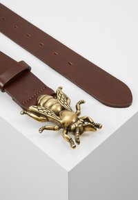 Brauner Ledergürtel mit einer großen, detaillierten Messingbiene als Schnalle, die aufwendige Flügel und Beine aufweist. Der Gürtel hat mehrere Löcher zur Anpassung.