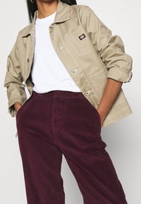 Veste beige en coton avec poches avant et boutons argentés, portée sur une chemise blanche et associée à un pantalon en velours côtelé bordeaux foncé.