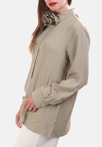 Camicia a maniche lunghe khaki con un accento floreale sul colletto, realizzata in tessuto liscio, con apertura frontale a bottoni e vestibilità rilassata.