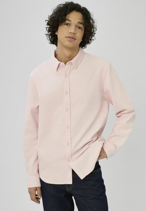 Chemise en velours côtelé rose clair avec col boutonné, manches longues et ourlet légèrement arrondi, portée avec un jean en denim foncé.