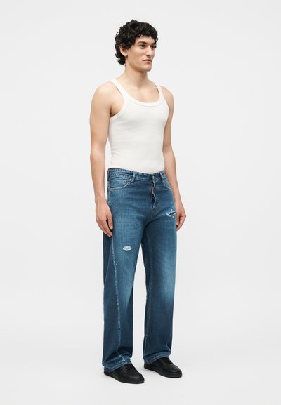 Νέος άνδρας με σγουρά μαλλιά που φοράει λευκό αμάνικο μπλουζάκι, φαρδιά μπλε τζιν με σκισμένες λεπτομέρειες και μαύρα αθλητικά παπούτσια, στέκεται με το πρόσωπο προς τα εμπρός.