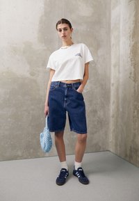 Hvit kort t-skjorte med logo, blå denimshorts, blå heklet veske og mørke sneakers med hvite detaljer; minimalistisk grå bakgrunn.