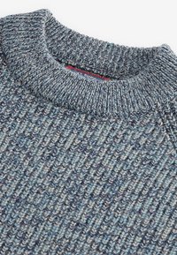 Primo piano di un colletto di un maglione a coste blu e grigio con cuciture testurizzate e un'etichetta rossa parzialmente visibile.