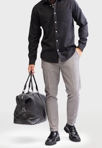 Black button-up shirt, gray plaid trousers, black leather shoes, carrying a large black leather duffel bag with silver hardware.