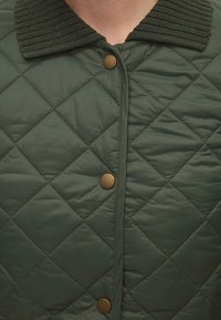 Gros plan d'une veste matelassée vert foncé avec des boutons-pression en bronze et un col côtelé, portée par une personne.