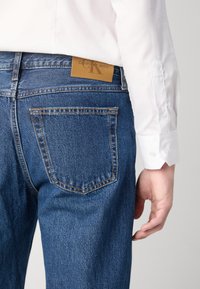 Jean en denim bleu avec une coupe classique, poches arrière et une étiquette en cuir beige avec le logo "Calvin Klein Jeans" sur la ceinture.