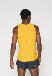 Yellow sleeveless athletic tank top with rounded neck, featuring a light texture and logo at the back. Paired with dark shorts.