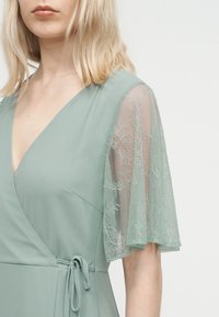 Ξανθιά γυναίκα που φοράει φόρεμα τύπου wrap σε φτέρη πράσινο χρώμα με μισά μανίκια από διαφανή δαντέλα και ζώνη που δένει στη μέση, μπροστά σε απλό φωτεινό φόντο.