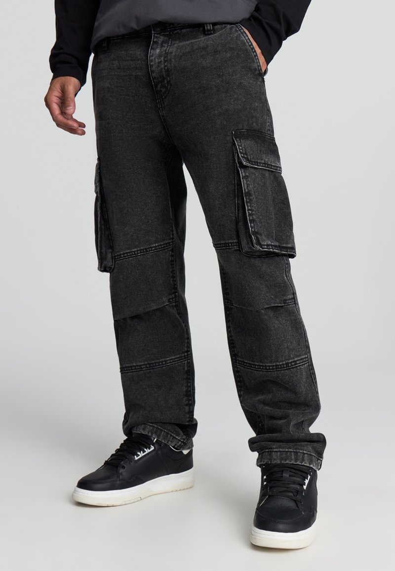 Czarne spodnie cargo z wieloma bocznymi kieszeniami, luźny krój i widoczne detale szwów. Noszone z czarnymi trampkami z białymi podeszwami.