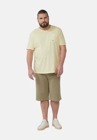 Hellgelbes Baumwoll-T-Shirt mit Brusttasche, kombiniert mit khakifarbigen Shorts bis zum Knie. Das Model trägt weiße Turnschuhe. Einfaches Design, lockere Passform.