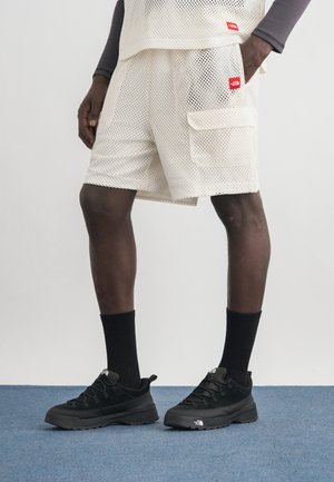 Persona che indossa pantaloncini bianchi a rete con tasca laterale e sneakers nere con marchio e calzini, in piedi su un pavimento con moquette blu.