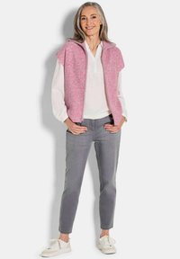 Gilet en tricot rose avec fermeture éclair sur le devant, porté sur une chemise blanche. Un jean gris slim et des baskets blanches complètent la tenue. Design et texture simples.
