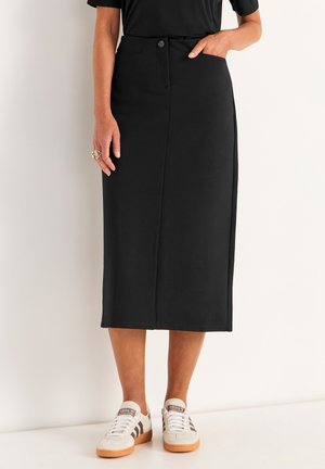 Femme portant une jupe midi noire avec des boutons sur le devant, un haut noir, des baskets blanches avec des rayures marron, et une bague en or à la main gauche.