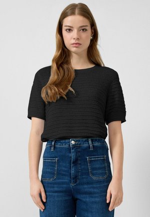 Młoda kobieta o długich brązowych włosach, ubrana w czarną, teksturowaną bluzkę z krótkim rękawem oraz niebieskie jeansy z wysokim stanem, stoi na tle jednolitego tła.