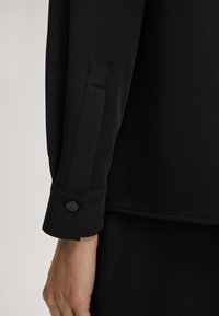 Zwarte blouse met lange mouwen, voorzien van een manchet met een zwarte knoop en subtiele textuur. De stof lijkt glad met een gestructureerde pasvorm.