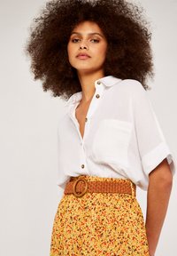 Weißes Leinenhemd mit kurzen Ärmeln und Knopfdetails, kombiniert mit einem gelben Blumenrock, der mit einem geflochtenen braunen Gürtel und einer runden Schnalle verziert ist.