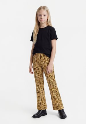 Ung jente med langt blondt hår iført svart t-skjorte, leopardmønstrede bukser og svarte ankelstøvler, stående mot en hvit bakgrunn.