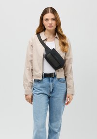 Svart läder crossbody-väska med lock, justerbar rem och detaljer i guld, bärs över en ljus jacka och vit skjorta.