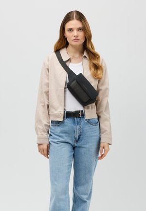 Borsa a tracolla in pelle nera con pattina, tracolla regolabile e dettagli in oro, indossata sopra una giacca leggera e una camicia bianca.