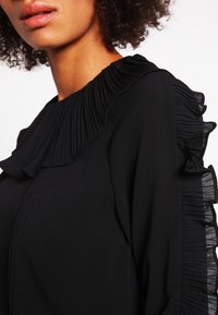 Blusa nera con un collo alto plissettato e maniche a strati con volant. Il tessuto appare liscio con una consistenza fluida.