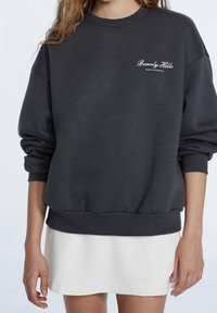 Sudadera gris oscura con cuello redondo, hombros caídos y puños acanalados. Presenta texto de logo blanco en la parte izquierda del pecho.