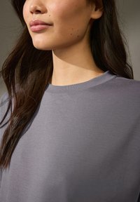 Grauer Pullover mit Rundhalsausschnitt und lässiger Passform; weicher Stoff mit glatter Textur; das Model hat langes, dunkles Haar.