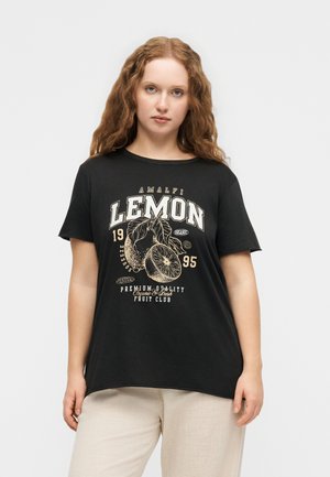 Femeie cu păr lung și ondulat, purtând un tricou negru cu un grafic vintage cu lămâi și pantaloni deschiși la culoare, stând în fața unui fundal simplu.