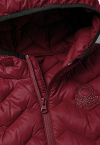 Bordó péřová bunda s kapucí, prošívaný vzor, přední zip a vyšívané logo. Obsahuje černý lem kolem otvoru kapuce.