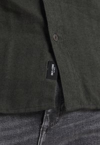 Camicia button-up a spina di pesce verde oliva con bottoni marroni e un'etichetta nera sull'orlo laterale. Il tessuto sembra texturizzato e robusto.