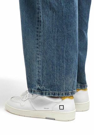 Sneaker bianche in pelle con dettagli perforati, accentuazione gialla sul tallone e suola testurizzata, abbinate a jeans larghi in denim blu.