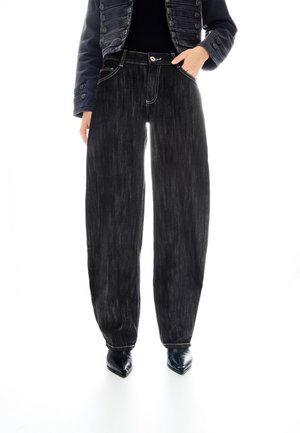 Osoba nosząca czarne jeansy o szerokich nogawkach, czarne szpiczaste buty oraz ciemną krótka kurtkę z ozdobnymi guzikami.