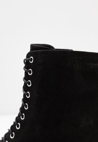 Stivaletto alla caviglia in camoscio nero con occhielli metallici e lacci neri, mostrato in una vista laterale ravvicinata su sfondo bianco.