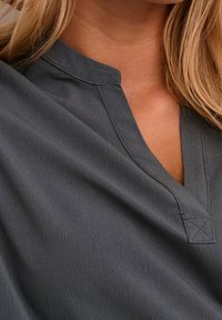 Nahaufnahme einer Frau, die eine strukturierte, dunkelgraue Bluse mit V-Ausschnitt und kleinem Stehkragen trägt, mit seitlich sichtbarem blonden Haar.