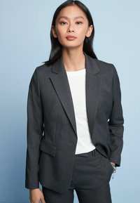Grå skræddersyet blazer med klapkrave og to forlommer, parret med en hvid t-shirt. Glat stof, tætsiddende silhuet, minimal detaljer.