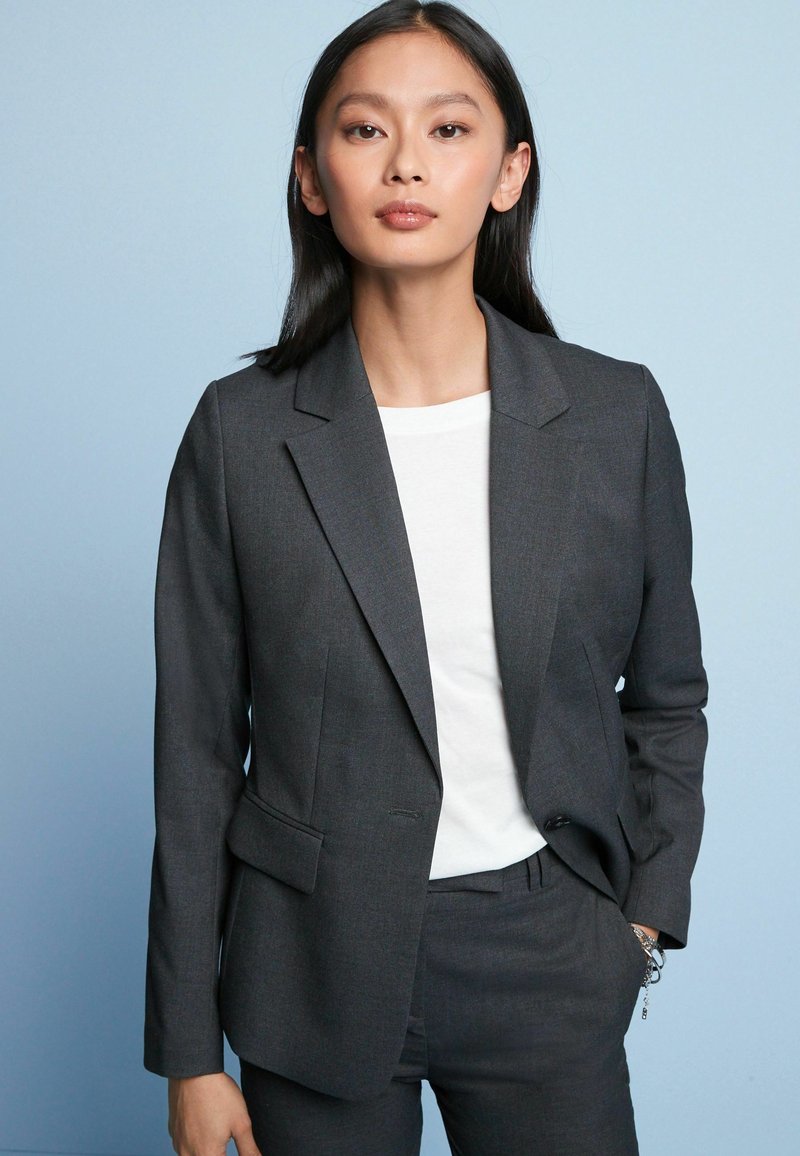 Grå skræddersyet blazer med klapkrave og to forlommer, parret med en hvid t-shirt. Glat stof, tætsiddende silhuet, minimal detaljer.