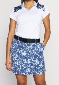 Valkoinen urheilupaita, jossa on siniset marmorikuvioiset hihansuut, yhdistettynä sinikuvioiseen hameeseen. Slim fit -leikkaus mustalla vyöllä.