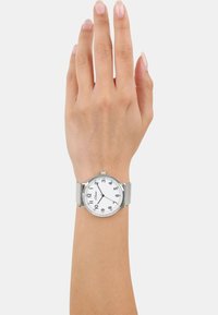 Silberne Uhr mit einem weißen Zifferblatt, schwarzen arabischen Ziffern und einem strukturierten Mesh-Armband. Auf einer hellhäutigen Hand präsentiert.