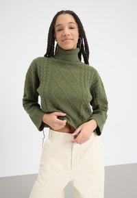 Pulover verde cu guler înalt tricotat, cu un design texturat și mâneci lejere, asortat cu pantaloni crem cu talie înaltă.