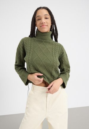 Groene kabelgebreide turtleneck trui met een textuurpatroon en losse mouwen, gecombineerd met crèmekleurige high-waisted broek.