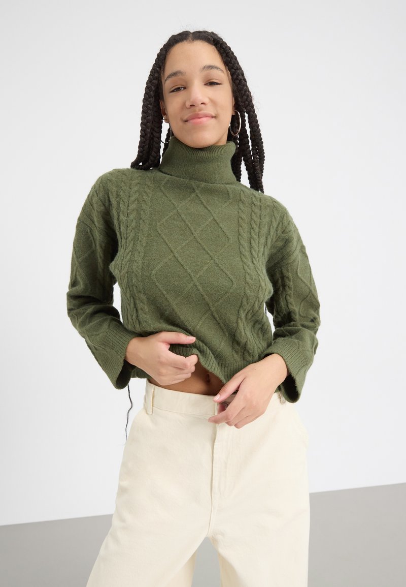 Pulover verde cu guler înalt tricotat, cu un design texturat și mâneci lejere, asortat cu pantaloni crem cu talie înaltă.