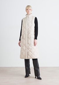 Colete longo acolchoado bege com colarinho alto, fecho à frente, dois bolsos de peste e cintura ajustável com cordão. Camisola e calças ajustadas pretas usadas por baixo.