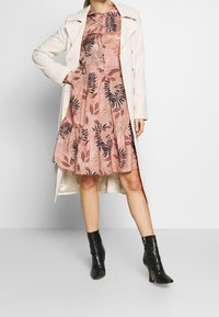 Robe florale rose clair avec des motifs de feuilles noirs, jupe à volants, associée à un manteau beige. Le mannequin porte des bottines à talons noires.